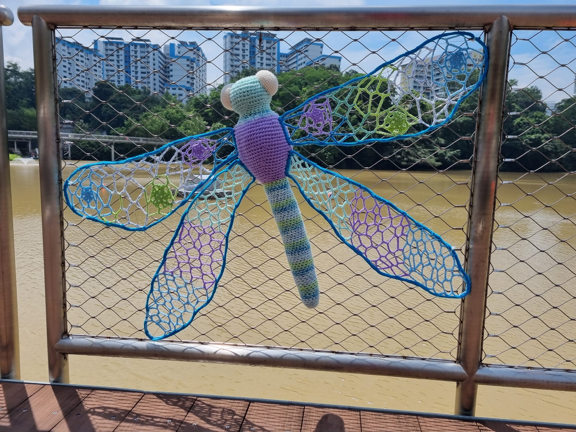 A crocheted dragonfly with lacy purple and teal net wings on the pond boardwalk railing.