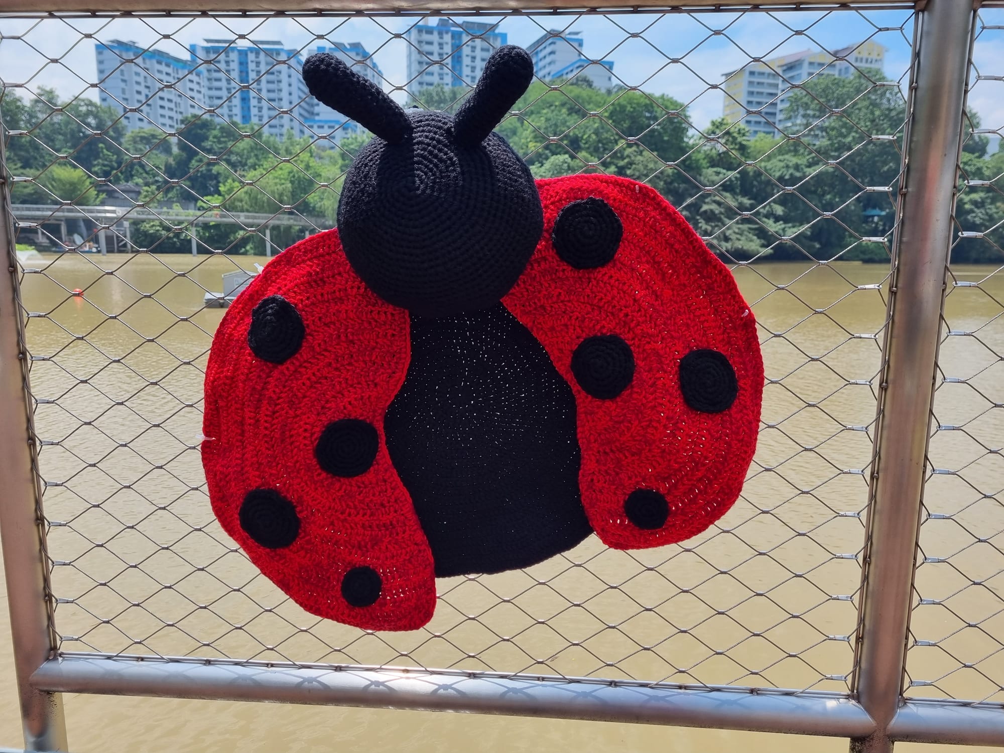 A large red and black crocheted ladybug with antennae on the pond railing.