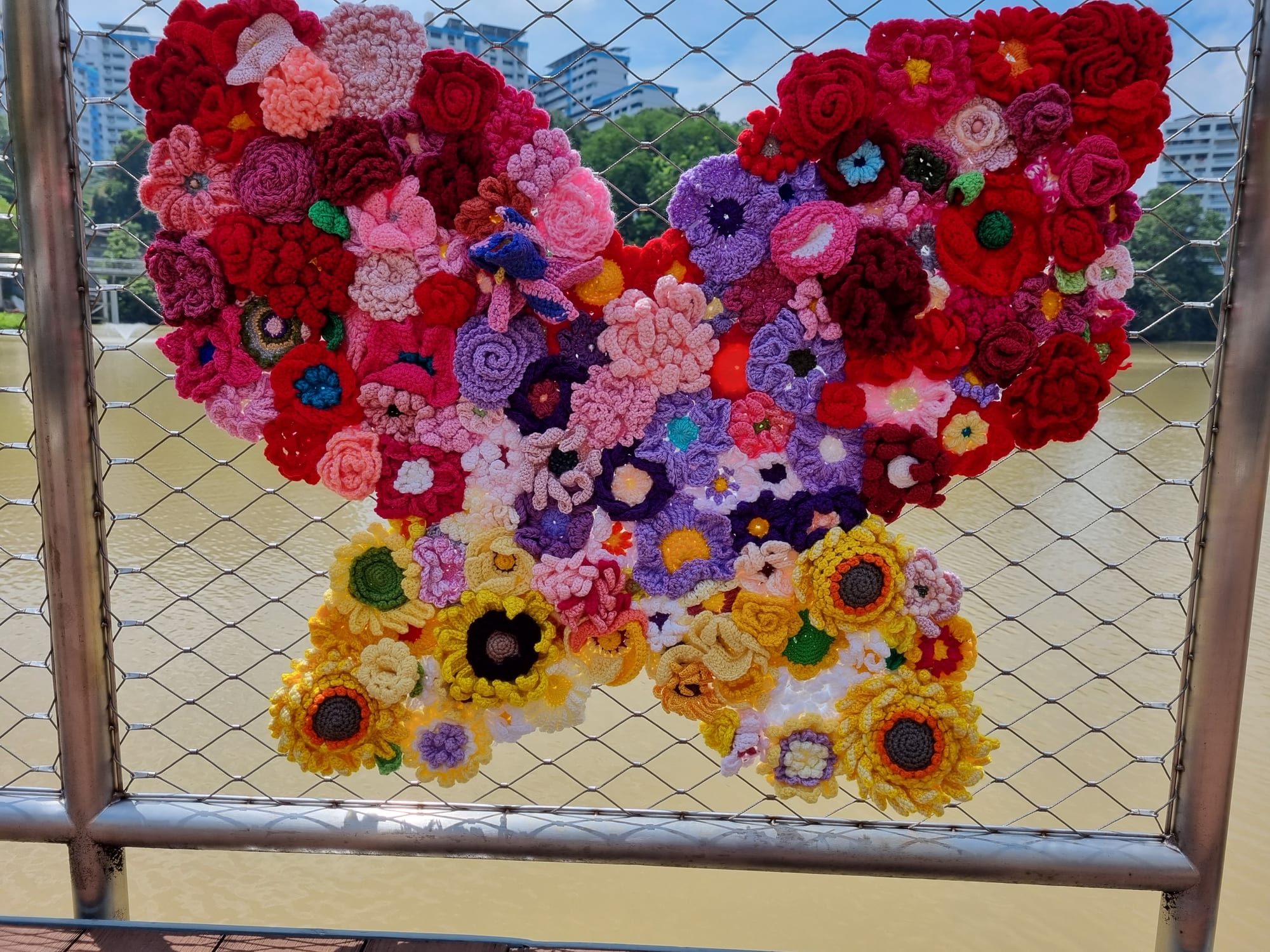 A crocheted butterfly of red and pink flowers with golden sunflower lower wings, on the cable railing of the Pangsua Pond boardwalk.