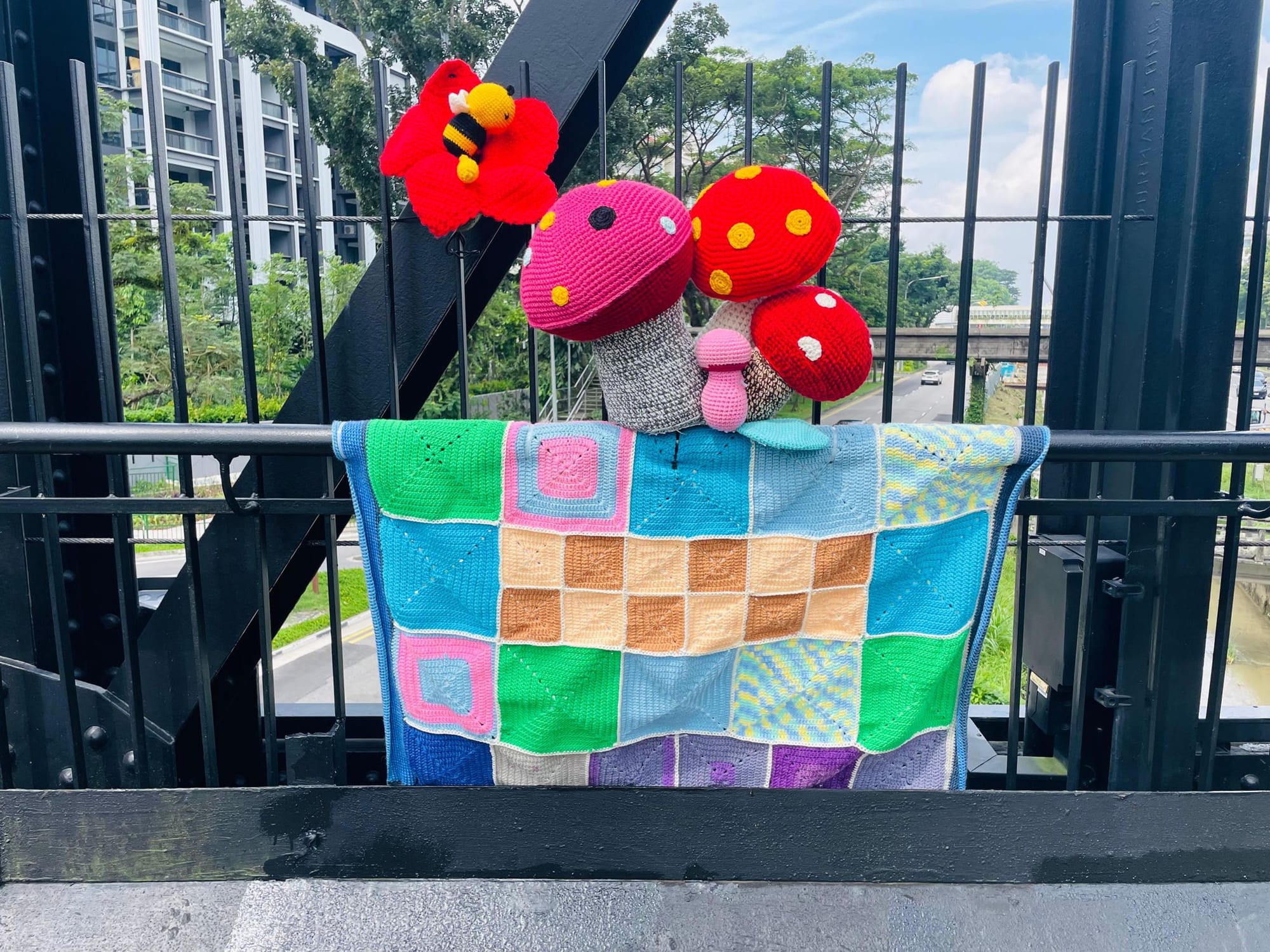 Crocheted toadstools and a bee above a checkered patchwork blanket on the bridge railing.